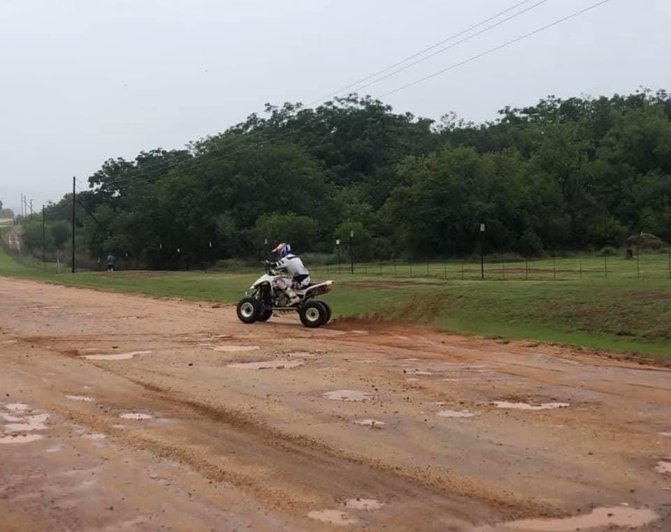 Two helmeted riders, one on a quad bike and one on a dirt bike, on a wet dirt road